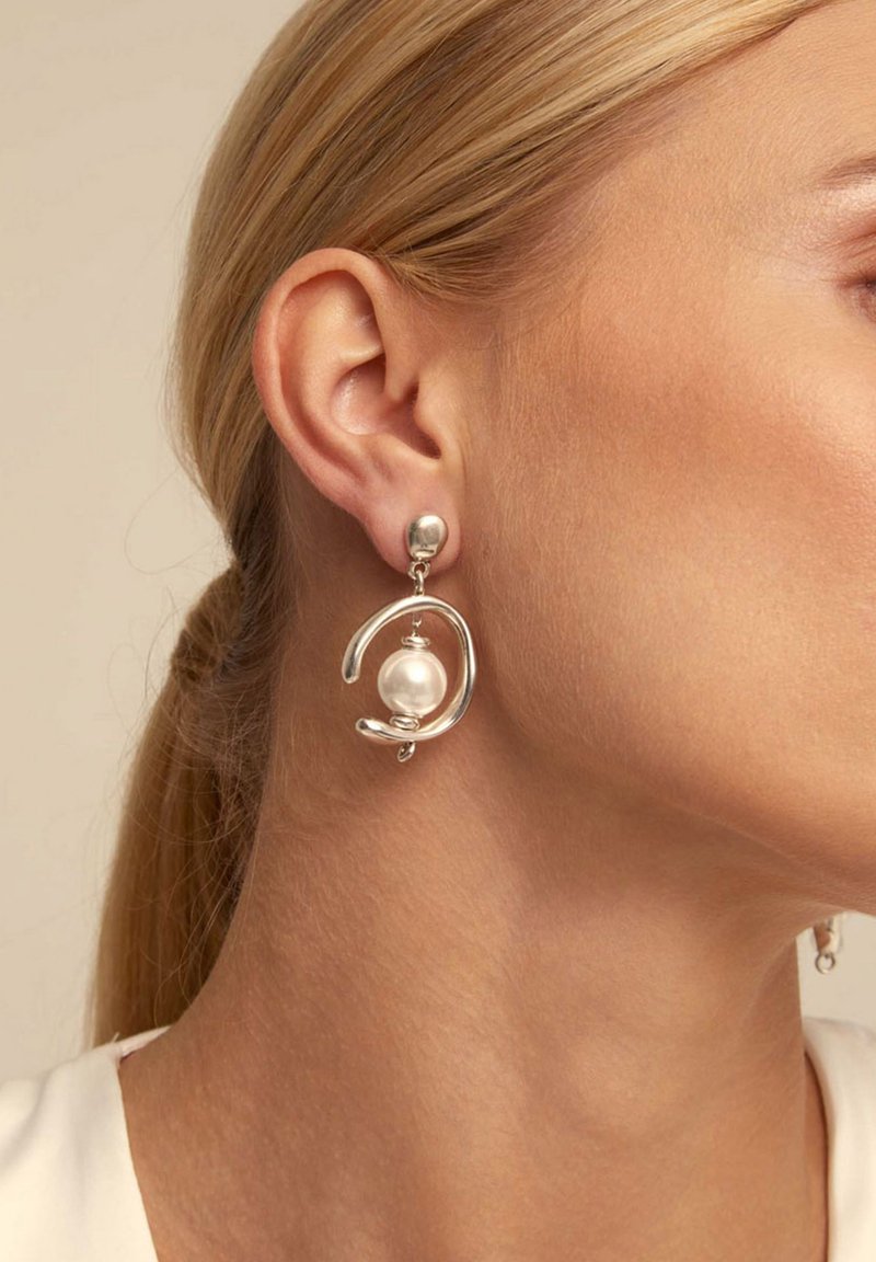 Mujer de piel clara y cabello rubio lleva un pendiente de plata con una gran perla blanca central y un diseño abstracto en forma de bucle.