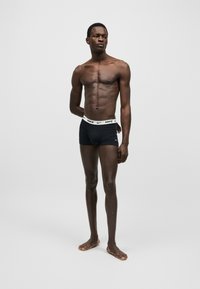 Λεπτός άνδρας με σκουρόχρωμο δέρμα, ξυπόλυτος, φοράει μαύρα ανδρικά εσώρουχα Nike με λευκή ζώνη, τα χέρια πίσω στην πλάτη, κοιτάει ελαφρώς αριστερά σε απλό γκρίζο φόντο.