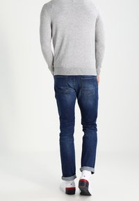 Grauer Pullover mit geripptem Saum, dunkelblaue Jeans, weiße Socken und weiße Schuhe mit roten Akzenten. Fokus auf die Rückansicht und lässige Passform.