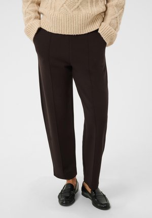 Personne portant un pantalon marron foncé ajusté, un pull en maille beige et des mocassins en cuir noir, debout les mains dans les poches.