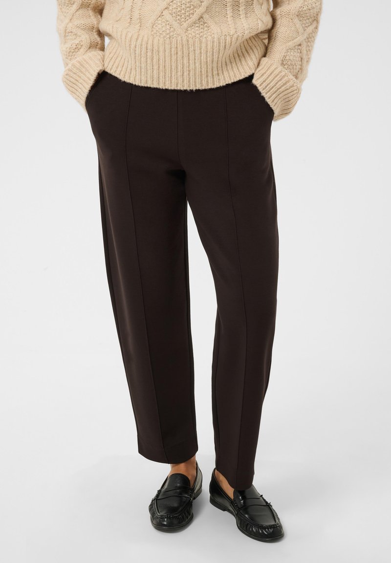 Personne portant un pantalon marron foncé ajusté, un pull en maille beige et des mocassins en cuir noir, debout les mains dans les poches.