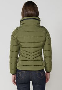 Chaqueta acolchada verde oliva con cuello alto, acolchado horizontal y en forma de Chevron, y detalles en negro en el escote y los puños.