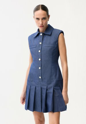 Kobieta w niebieskiej sukience bez rękawów z srebrnymi guzikami i plisowaną spódnicą, stojąca na tle jednolitego białego tła.