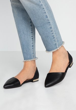 Ballerines noires à bout pointu avec un dos texturé. Le talon bas présente un accent doré. Associées à un jean en denim avec un ourlet effiloché.