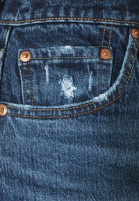 Acercamiento al pequeño bolsillo delantero de unos jeans de mezclilla azul con costuras naranjas y remaches de cobre, mostrando detalles de tejido desgastado y deshilachado.