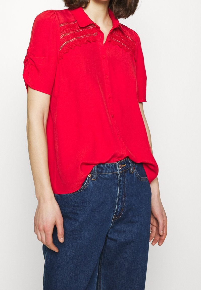 Camicia rossa con bottoni, presenta maniche corte, dettagli in pizzo sullo scollo e una vestibilità rilassata. Abbinata a jeans in denim blu scuro.