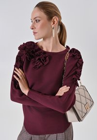 Maglione bordeaux con accentuazioni arruffate sulle spalle, abbinato a una borsa beige trapuntata con chiusura dorata e tracolla a catena.