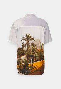 Chemise à manches courtes boutonnée avec un imprimé paysage tropical comprenant des palmiers, des plantes, des bâtiments et une grande colline rocheuse en arrière-plan.