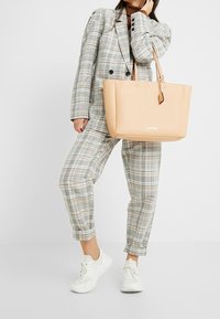 Tan tote bag met een gladde, matte textuur en een minimalistisch ontwerp. Beschikt over twee stevige handvatten en een subtiele logo aan de voorkant.