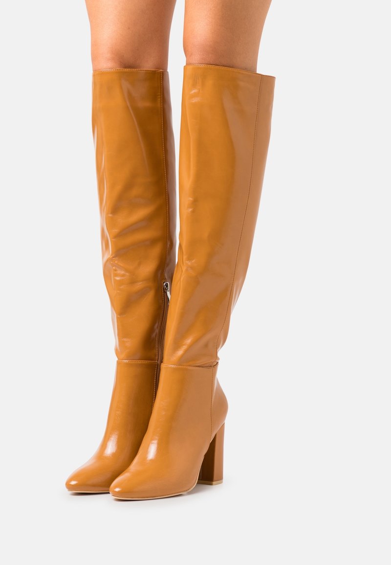 Botas de cuero caramelo brillantes hasta la rodilla, con puntera afilada y tacón grueso. Detalle de cremallera lateral y textura suave.