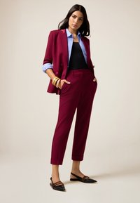 Bordeaux-Blazer und -Hose mit einem hellblauen Hemd darunter. Schwarze Ballerinas mit dekorativen Akzenten. Glattes Material und taillierte Passform.