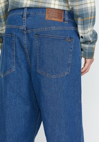 Jeans de mezclilla azul con un diseño de pierna ancha, parche de cuero marrón en la cinturilla y dos bolsillos traseros con costuras en contraste.