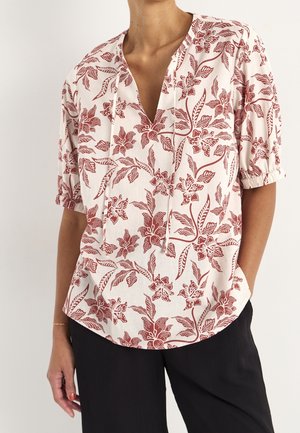 Kobieta w kremowej bluzce w czerwony kwiatowy wzór, z półdługimi rękawami, dekoltem w serek z wiązaniami, w połączeniu z czarnymi spodniami.