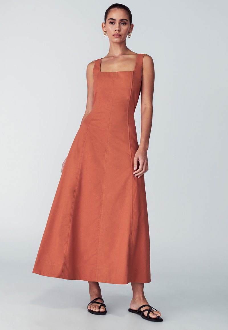 Maxi šaty bez rukávov v farbe hrdzavej s štvorcovým výstrihom a áčkovým strihom. Vyrobené z textúrovaného materiálu s vertikálnymi švami. Čierne sandále.