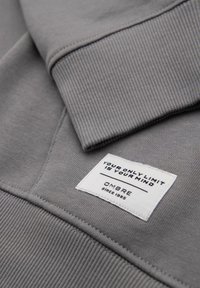 Szary sweatshirt z ribbowanym kołnierzem i mankietami. Zawiera białą metkę z czarnym tekstem, wyszytą na materiale. Miękka tekstura, minimalistyczny design.