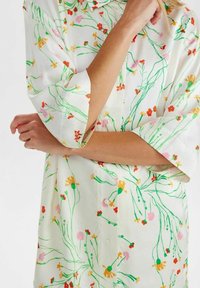 Femme portant un chemisier crème boutonné avec des manches retroussées et un motif floral coloré dans des tons de vert, rouge, rose et jaune.