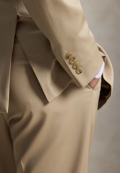 Persona che indossa un abito beige con quattro bottoni sulla manica e la mano posata casualmente in tasca.
