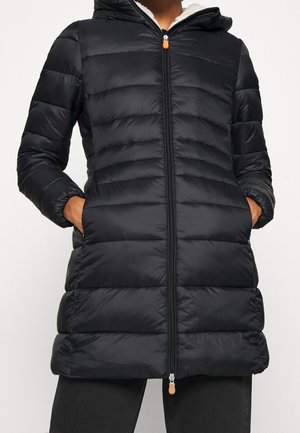 Osoba ubrana w długi czarny puchowy płaszcz z kapturem, z przodu na zamek błyskawiczny, z rękami w bocznych kieszeniach, stojąca na jednolitym tle.