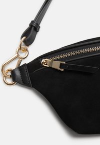 Bolso de ante negro con cremallera y herrajes dorados. Cuenta con una correa de cuero y un detalle de cierre, mostrando una textura suave y una forma angular.