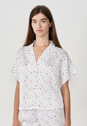 Jeune femme portant une chemise blanche à boutons et à manches courtes ainsi que des shorts assortis avec un petit motif de cœurs roses et bleu marine.