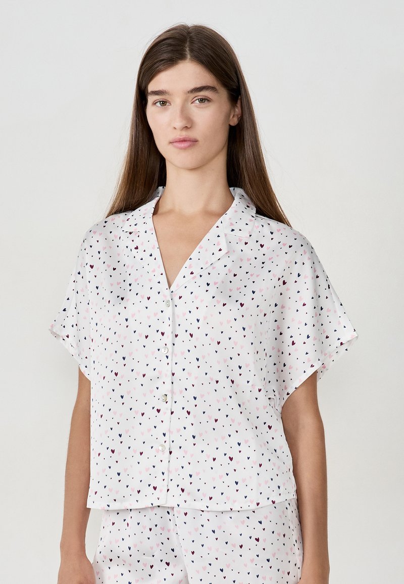 Jonge vrouw in een wit overhemd met korte mouwen en bijpassende shorts met een klein roze en marineblauw hartpatroon.