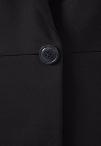 Gros plan d'un manteau noir avec un seul bouton noir brillant à quatre trous fixant deux panneaux de tissu qui se chevauchent.
