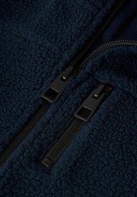 Námořnická fleecová bunda se strukturovaným povrchem, dvojitými černými zipy a diskrétní značkovou nášivkou poblíž zipu.