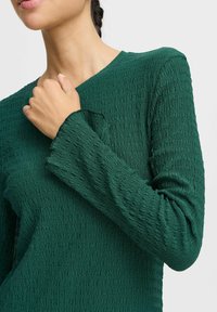 Haut long à manches vert texturé avec un col rond et des détails de poignets effilochés, mettant en avant un motif de tissu froissé.