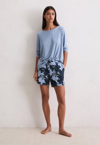 Kobieta stojąca boso przy białej ścianie, ubrana w jasnoniebieską bluzkę z długim rękawem i niebieskie spodenki w kwiatowy wzór, z rękami w kieszeniach.