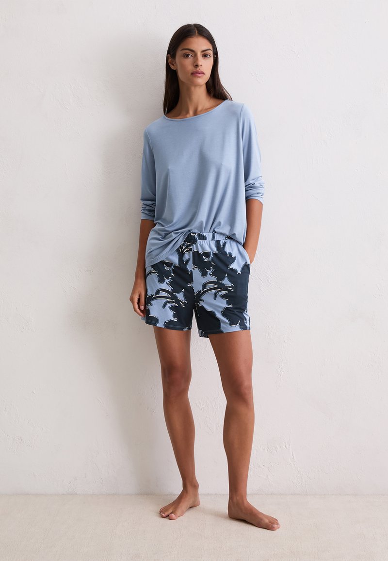 Kobieta stojąca boso przy białej ścianie, ubrana w jasnoniebieską bluzkę z długim rękawem i niebieskie spodenki w kwiatowy wzór, z rękami w kieszeniach.