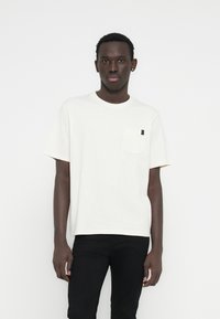 Λευκό μπλουζάκι με κοντά μανίκια, μικρό θηλυκό τσέπη στο στήθος και μαύρη ετικέτα ως διακοσμητικό στοιχείο. Το υλικό φαίνεται μαλακό και ελαφρώς υφασμένο.