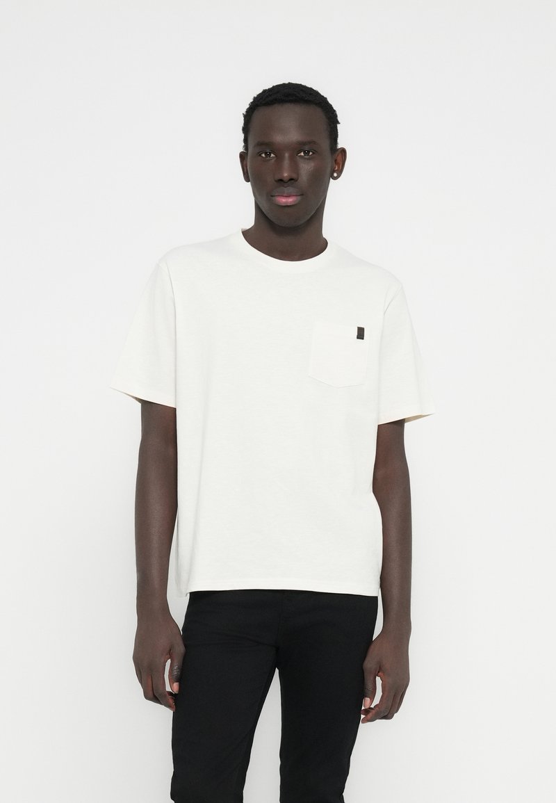 Λευκό μπλουζάκι με κοντά μανίκια, μικρό θηλυκό τσέπη στο στήθος και μαύρη ετικέτα ως διακοσμητικό στοιχείο. Το υλικό φαίνεται μαλακό και ελαφρώς υφασμένο.