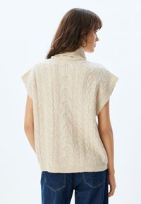 Beige gebreide trui met een kabeldesign, hoge hals en korte mouwen, weergegeven vanuit een achteraanzicht tegen een lichte achtergrond.