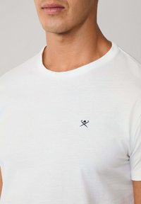 Bílý bavlněný tričko s kulatým výstřihem, doplněné malým tmavě modrým vyšívaným vzorem překřížených klacků na levé straně hrudi.