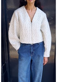 Hvid tekstureret jakke med lynlås foran, featuring en v-hals og voluminøse ærmer, båret over blå denimjeans.