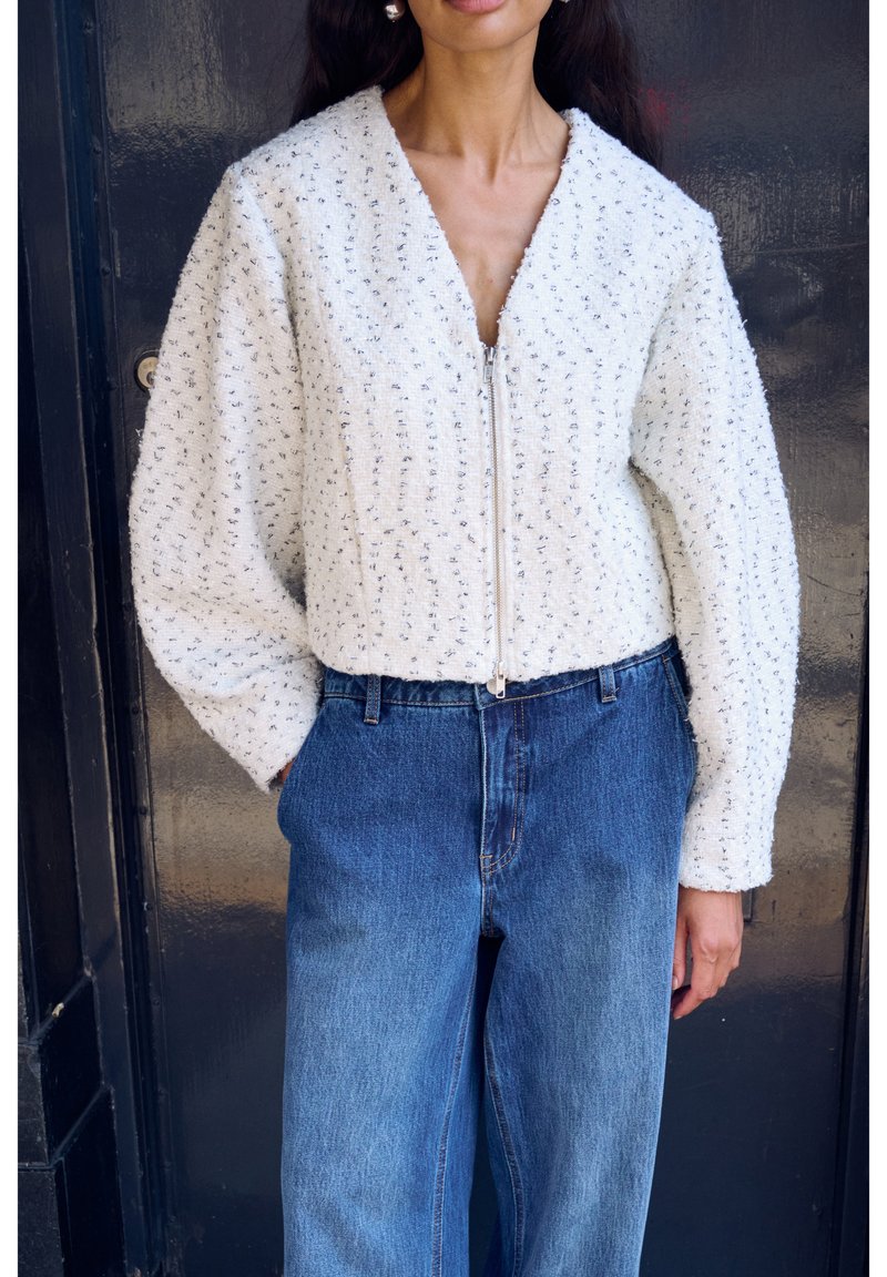 Hvid tekstureret jakke med lynlås foran, featuring en v-hals og voluminøse ærmer, båret over blå denimjeans.