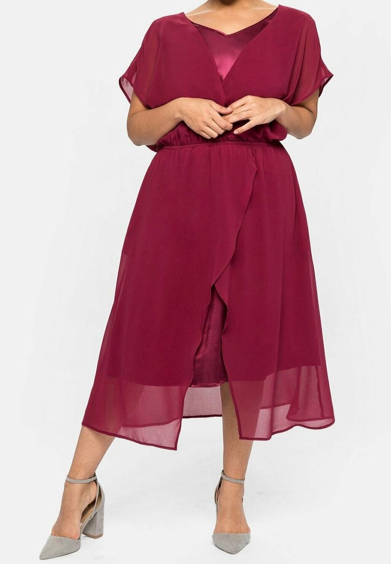 Robe bordeaux mi-longue en tissu léger, présentant un design en portefeuille, des manches courtes et une taille cintrée. Accentuée par un look superposé.