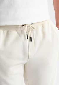 Pantalones de chándal color crema con cintura elástica, cordón con extremos negros y bolsillos laterales. Textura suave, ajuste casual.