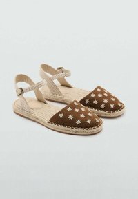 Sandales en tissu marron avec broderie de fleurs blanches. Des brides de cheville beige texturées et des semelles en jute complètent le design.