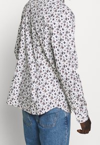 Bílá košile s dlouhým rukávem se květinovým vzorem v tmavě modré, oranžové a zelené barvě. Materiál má jemnou texturu a volný střih.