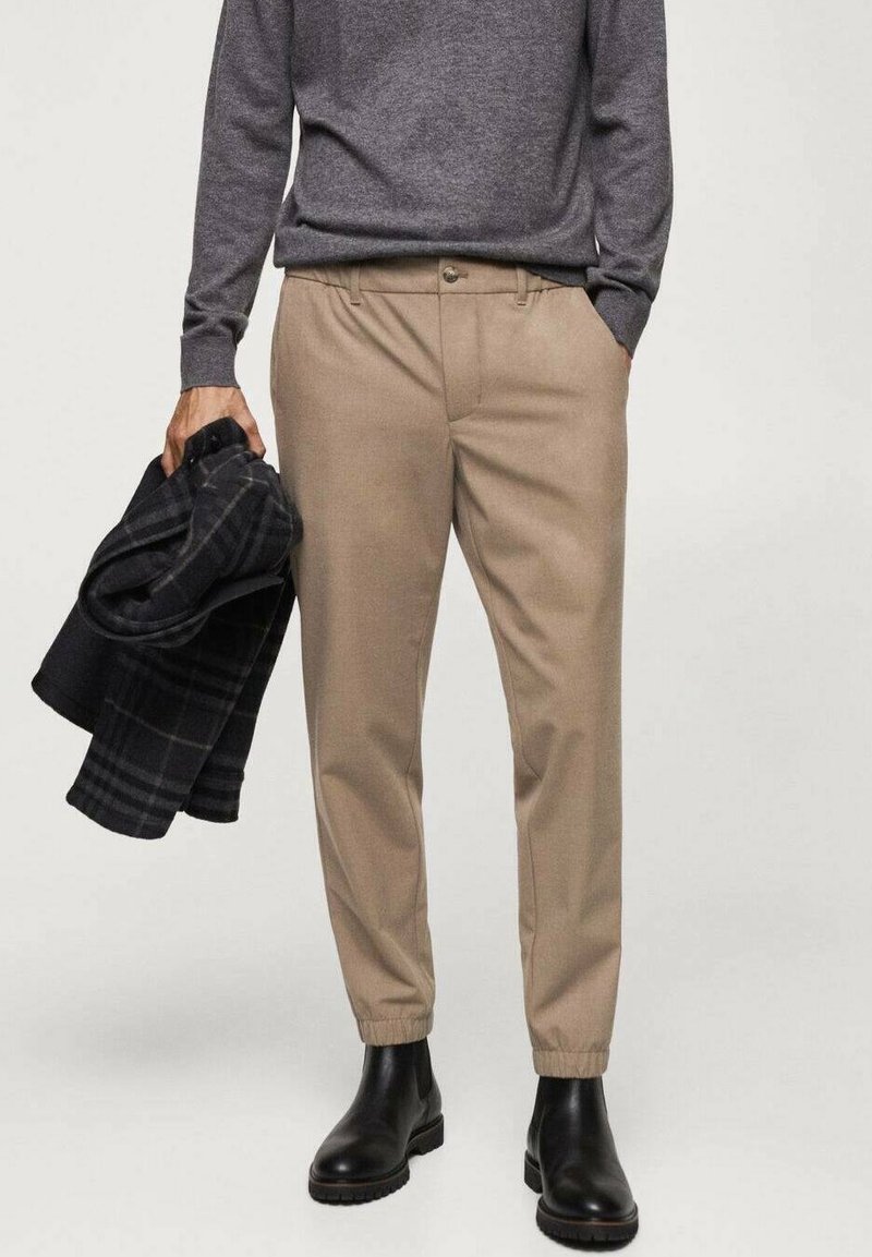 Personne portant un pantalon beige fuselé avec des poignets élastiques, des bottes noires, un pull gris, tenant un manteau noir à carreaux plié dans une main.