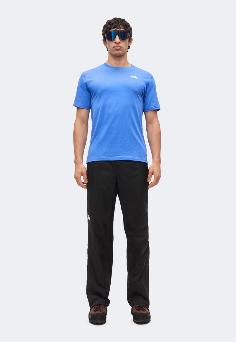 Hombre con una camisa azul de manga corta, pantalones negros, zapatos de senderismo marrones y grandes gafas de sol reflectantes de pie mirando hacia adelante.