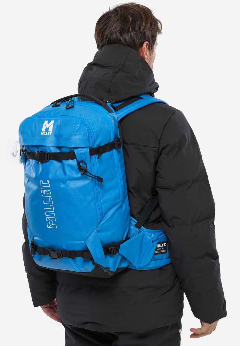 Un sac à dos bleu en tissu texturé et durable, avec des bretelles réglables, des boucles de compression et un logo sur le devant. Porté sur une veste noire.