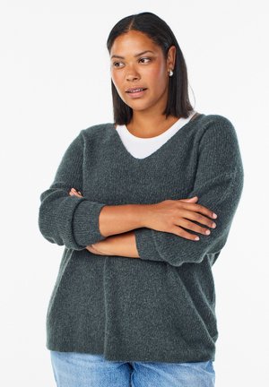 Donna dalla carnagione media e capelli lisci indossa un maglione grigio sopra una camicia bianca, con le braccia incrociate e lo sguardo rivolto di lato, su uno sfondo bianco.