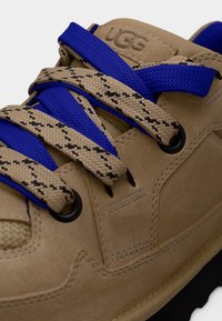 Baskets en daim camel avec des lacets à motifs bleu et beige passant par des œillets noirs, ornées du logo UGG sur la languette.