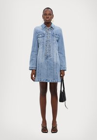 Denimkjole i lyseblå farge, lange ermer, snøring i front, krage og to brystlommer. Par med svarte sandaler med stropper.