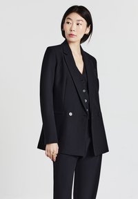 Donna che indossa un blazer nero su misura, con un gilet e pantaloni coordinati, che guarda a sinistra su uno sfondo grigio uniforme.