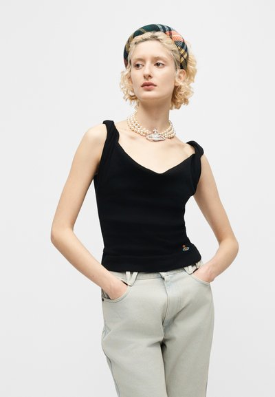 Νεαρή γυναίκα που φοράει μαύρο αμάνικο τοπ, ανοιχτόχρωμο τζιν, κολιέ με μαργαριτάρια και σχέδιο κορδέλα στο κεφάλι, στέκεται με τα χέρια στις τσέπες μπροστά σε λευκό φόντο.