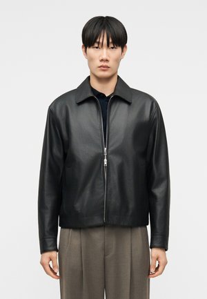 Jeune homme portant une veste en cuir noire zippée et un pantalon taupe plissé, debout devant un fond clair uni.