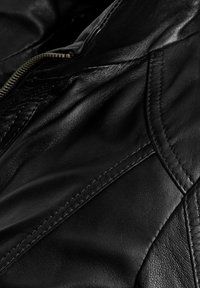 Crna kožna jakna s glatkom teksturom, vidljivim šavovima i kutnim dizajnom. Ima zatvaranje na patentni zatvarač i suptilan sjaj.